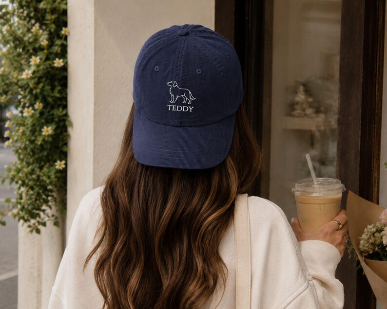May include: A navy blue baseball cap featuring an embroidered white line art illustration of a dog above the text TEDDY in a simple white font. The cap has a structured crown, a curved brim, and a button at the top centre of the fabric panels.
