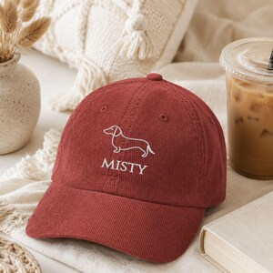 May include: A dark red corduroy baseball cap featuring an embroidered white line art illustration of a dachshund dog above the text MISTY in a white serif font. The cap has a structured crown, a curved brim, and stitched eyelet details.