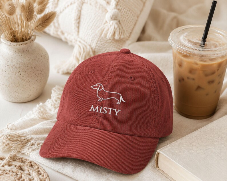 May include: A dark red corduroy baseball cap featuring an embroidered white line art illustration of a dachshund dog above the text MISTY in a white serif font. The cap has a structured crown, a curved brim, and stitched eyelet details.