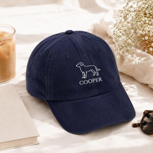 May include: A navy blue corduroy baseball cap featuring an embroidered white line art illustration of a dog above the text COOPER. The cap has a structured crown, a curved brim, and stitched eyelets on the panels for ventilation and durability.