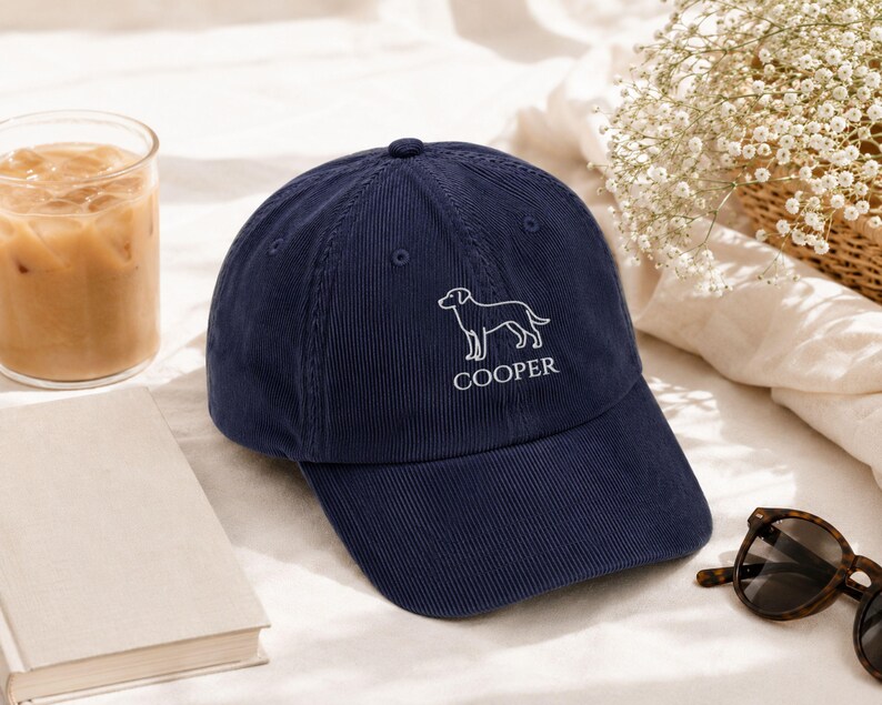 May include: A navy blue corduroy baseball cap featuring an embroidered white line art illustration of a dog above the text COOPER. The cap has a structured crown, a curved brim, and stitched eyelets on the panels for ventilation and durability.