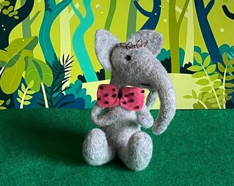 May include: A gray felt elephant toy wearing a pink bow tie with white polka dots. The elephant is sitting on a green surface in front of a green and blue cartoon forest background.