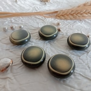 Puede incluir: Cinco botones redondos y planos con un esquema de color verde y crema. Los botones tienen un diseño de círculo concéntrico elevado.