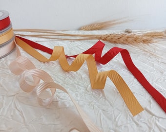 May include: Assortment of ribbon spools in beige, gold, and red colors. The ribbon is curled and arranged on a white textured surface.