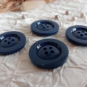 May include: Four navy blue, round buttons with four holes each are arranged on a white, textured surface. The buttons have a glossy finish and a raised center. Decorative elements are in the background.