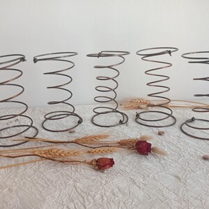 May include: Five antique metal springs, each with a coiled design and a circular top and bottom. The springs are a dark bronze color and are arranged in a row. Dried wheat stalks and red roses are placed in front of the springs.
