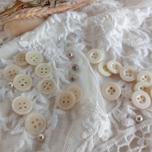 Può includere: Una collezione di bottoni bianchi perlacei con quattro fori ciascuno, disposti su uno sfondo di tessuto in pizzo bianco.