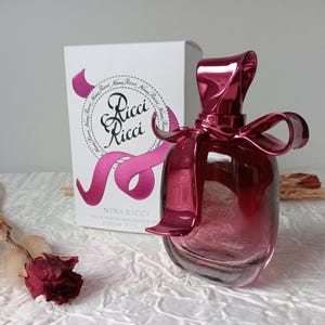 May include: A Nina Ricci perfume bottle with a shiny, deep pink bow-shaped top and a matching box. The bottle is a gradient of pink and clear glass. The box has the words "Ricci Ricci" and a pink ribbon design.