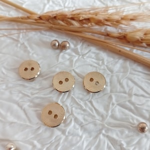 Puede incluir: Cuatro botones de color dorado con dos agujeros cada uno. Los botones tienen una superficie texturizada y están dispuestos sobre un fondo blanco.