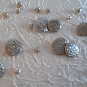 May include: Six silver buttons with a textured surface, scattered on a white fabric background.