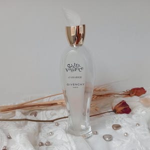 May include: A clear glass perfume bottle with a gold cap and a white feather-like accent. The bottle has the text "Extra Vagance d'Amarige GIVENCHY Paris". Dried flowers and pearls are scattered around the bottle.