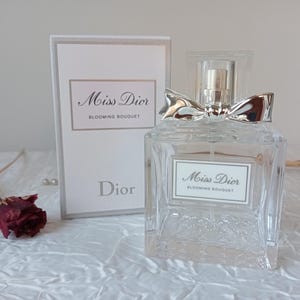 Può includere: Un flacone di profumo in vetro trasparente con un fiocco argentato sulla parte superiore e un'etichetta bianca con la scritta "Miss Dior Blooming Bouquet". Una scatola abbinata è dietro la bottiglia. Una rosa rossa secca e una spiga di grano sono in primo piano.