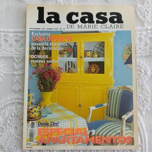 Op de afbeelding: Een vintage La Casa de Marie Claire tijdschrift van december 1994 tot januari 1995, met een felgele kast, een blauw-wit gestreepte fauteuil, een gele vaas met roze bloemen en een geruit tafelkleed op de omslag met opvallende tekst.