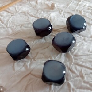 May include: Five dark gray, faceted beads with a glossy finish are scattered on a white, textured surface. The beads have a rounded, square shape and a two-tone color scheme, with a lighter gray top and a darker gray base. The beads are likely for jewelry making.