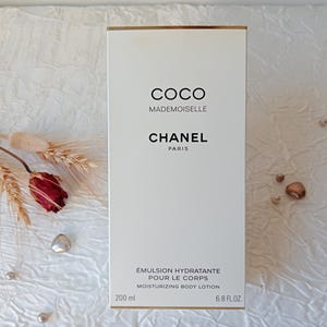 Può includere: Una scatola bianca di lozione per il corpo idratante Chanel Coco Mademoiselle. La scatola ha finiture dorate e testo nero che recita "COCO MADEMOISELLE CHANEL PARIS". La lozione è da 200 ml / 6,8 fl oz. Fiori secchi e perline decorative sullo sfondo.