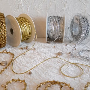 May include: An assortment of decorative ribbons and cords in gold and silver tones. The image shows spools of metallic cord and beaded trim, along with loose strands and embellishments. The items are arranged on a white, textured surface.