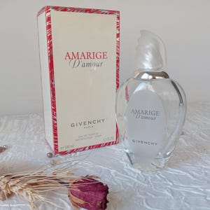 May include: A clear glass perfume bottle and box set. The bottle has a unique shape with a silver cap and the words "Amarige D'amour" and "Givenchy" printed on the front. The box is cream-coloured with red accents and the same text.