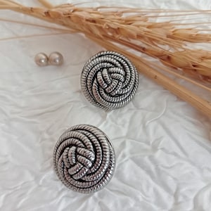 Puede incluir: Un par de pendientes de plata con un diseño anudado. Los pendientes son redondos y tienen una superficie texturizada.