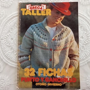 May include: A spiral-bound instructional booklet titled GRECA TALLER 32 FICHAS PUNTO Y GANCHILLO OTOÑO-INVIERNO. The cover features a photograph of a person wearing a grey knit cardigan with a multi-colored geometric pattern and red buttons.
