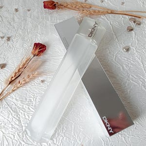 May include: A rectangular perfume bottle and its silver box are displayed on a white, textured surface. The bottle is clear with a frosted appearance, and the box has the brand name "DKNY" printed on it. Dried wheat stalks and a red rose are also present.