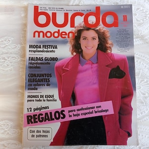 May include: A vintage issue of Burda Moden magazine, featuring a woman in a pink blazer and a pink shirt. The magazine cover has the title "Burda Moden" in large red letters, with text in Spanish. The cover also includes the number "11".