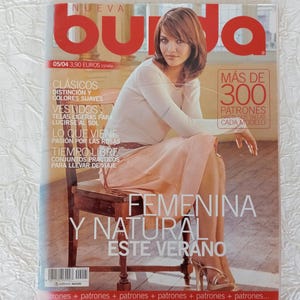 Peut inclure: Une couverture de magazine avec une femme en jupe rose clair et haut blanc, assise sur une chaise en bois. Le titre du magazine est "Nueva Burda" en rouge. Le texte de la couverture comprend "Más de 300 Patrones" et "Femenina y Natural Este Verano."