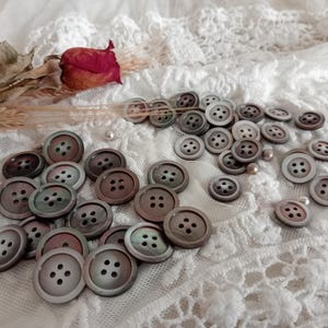 Peut inclure: Une collection de boutons ronds, irisés, en nacre dans des tons de gris, brun et vert. Les boutons sont disposés sur un fond de tissu en dentelle blanche.