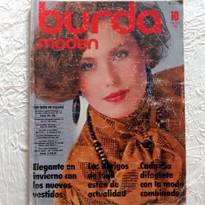 Puede incluir: Portada de la revista Burda Moden de octubre de 1990. Presenta a una mujer con cabello castaño y labios rojos. La portada incluye texto en español y titulares de moda. La revista está sobre una superficie blanca texturizada.