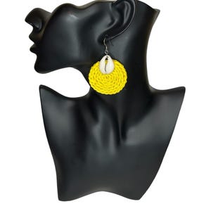 May include: Yellow woven circle earrings with a white seashell accent. The earrings are hanging from a mannequin's ear.