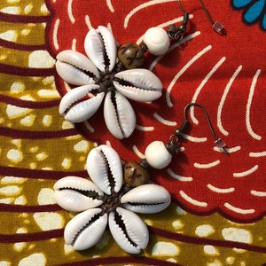 May include: A pair of earrings made with white seashells, brown beads, and a white bead. The earrings are hanging from a brown fabric background with a colorful pattern.