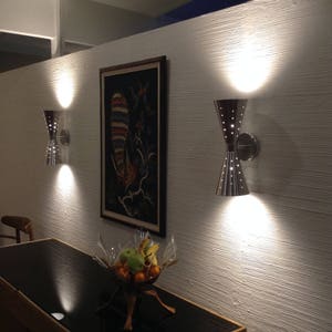 May include: Two silver wall sconces with a modern design illuminate a white wall with a textured finish. A framed painting with a colorful bird design hangs on the wall between the sconces. A bowl of fruit sits on a black table in the foreground.