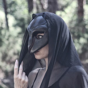 May include: A person wearing a black mask and a black hooded cloak, standing in a forest. The mask has a bird-like design. The person is wearing a black top with lace detailing. The background is blurred with green trees.