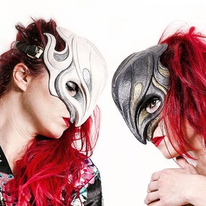 May include: Two people wearing decorative masks with flame-like designs. One mask is white, the other is black and gold. Both have red hair and are wearing patterned robes. The background is white.