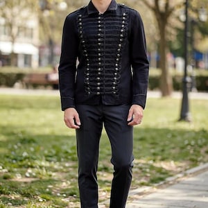 Peut inclure: Veste noire de style militaire ornée de boutons et de garnitures argentés. La veste a un col montant et est portée avec un pantalon noir et des chaussures beiges. La personne se tient sur une allée pavée dans un parc.