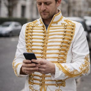 Peut inclure: Veste blanche ornée de détails dorés, notamment des boutons et des bordures décoratives. La veste a un col montant et est portée par une personne tenant un smartphone. Les détails dorés sont une caractéristique importante.