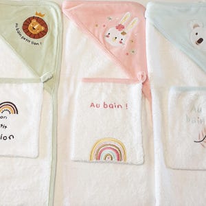 Peut inclure: Trois serviettes de bain à capuche blanches pour bébés avec des motifs brodés. L'une représente un lion, une autre un lapin et la troisième un koala. Chaque serviette a une poche avec du texte brodé et un motif arc-en-ciel ou feuille.