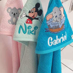 Peut inclure: Trois serviettes en tissu éponge suspendues, de couleurs rose, vert clair et bleu vif. La serviette rose présente un koala, la verte montre Mickey Mouse avec le nom Niki, et la bleue affiche Dumbo avec le nom Gabriel brodé dessus.