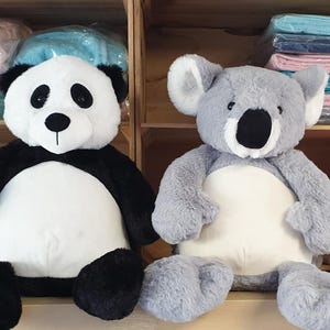 Peut inclure: Une peluche panda avec une fourrure noire et blanche et un grand ventre blanc est assise à côté d'une peluche koala avec une fourrure grise, des oreilles intérieures blanches et un grand ventre crème. Une bobine de fil gris est posée à droite.