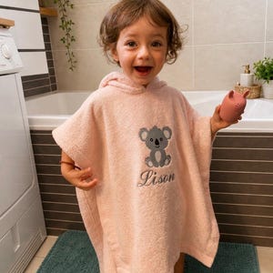 Peut inclure: Un poncho de bain à capuche rose clair orné d'une broderie représentant un koala gris avec le prénom Lison cousu en fil gris foncé en dessous. Le vêtement est fabriqué en tissu éponge doux et comprend une capuche pour plus de confort.