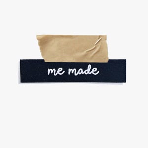 May include: A black rectangular label with the words "me made" in white script is adhered to a white background with a piece of brown tape. The tape is torn and partially covers the label.