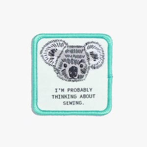 May include: Square patch with a teal border and a koala bear design. The koala is in black and white, with the text "I'M PROBABLY THINKING ABOUT SEWING." in a sans-serif font. The patch is likely for sewing projects.