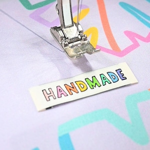 May include: A close-up of a sewing machine needle and foot stitching a white fabric with colorful abstract shapes. A rectangular label with the word "HANDMADE" in rainbow colors is positioned below the needle.