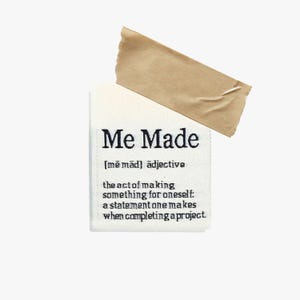 May include: A white rectangular label with the text "Me Made" in black, along with a definition. A piece of brown tape is adhered to the top of the label. The label is on a white background.