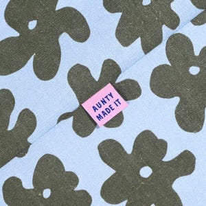 May include: Close-up of a light blue fabric with a repeating pattern of dark green floral shapes. A small pink tag with the text "AUNTY MADE IT" is sewn onto the fabric. The fabric has a soft, textured appearance.