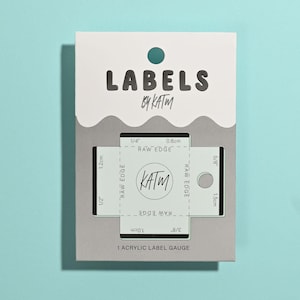 May include: A white and gray rectangular card labeled LABELS BY KATM containing a clear acrylic label gauge. The tool features various measurements in inches and centimeters, including 1/4 inch, 0.6cm, 1/2 inch, 1.2cm, 3/8 inch, 1.0cm, 5/8 inch, and 1.5cm.