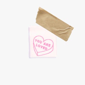 May include: A white patch with a pink heart-shaped candy design and the words "YOU ARE LOVED" embroidered on it. A piece of brown tape is adhered to the top of the patch.