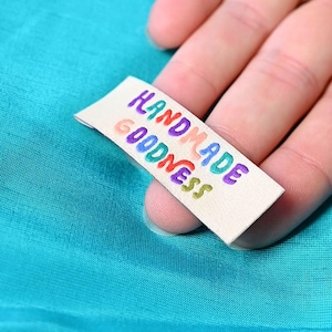 May include: A rectangular off-white fabric label featuring the embroidered text "HANDMADE GOODNESS" in multi-colored letters, including purple, red, orange, blue, and green, positioned against a solid bright turquoise fabric background.
