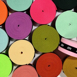 May include: A collection of colorful spools of webbing in various shades, including pink, green, orange, and purple. One pink spool has a black band with the text "KATIM". The spools are arranged closely together, creating a vibrant, textured pattern.