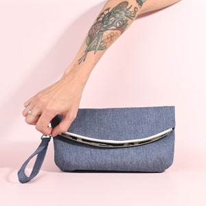 May include: A denim clutch with a white zipper and a black and white patterned lining. The clutch has a short strap.