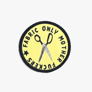 May include: A round yellow patch with black text and a black border. The text reads "FABRIC ONLY MOTHER FUCKERS" around the edge and "ONLY" above a pair of crossed scissors. A black star is on the left side.